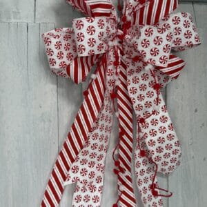Red and white peppermint Christmas bow with glitter candy cane striped ribbon, peppermint candy patterns, pom pom accents, and long flowing tails.