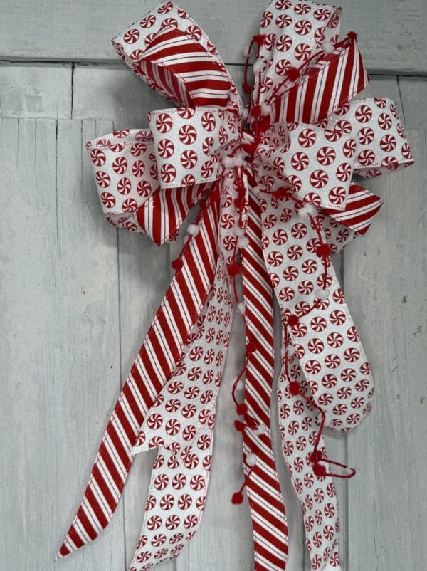 Red and white peppermint Christmas bow with glitter candy cane striped ribbon, peppermint candy patterns, pom pom accents, and long flowing tails.